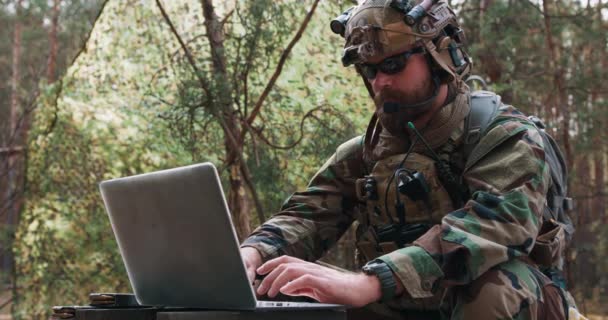 Retrato de comandante barbudo em um uniforme militar em um colete tático com um capacete na cabeça digitando no teclado de um laptop em uma base florestal temporária — Vídeo de Stock