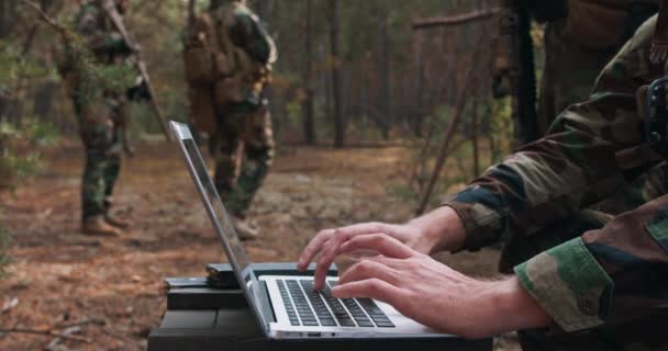 Portret van een bebaarde commandant in een militair uniform in een tactisch vest met een helm op zijn hoofd typend op het toetsenbord van een laptop in een tijdelijke bosbasis Op de achtergrond zie je een soldaat — Stockvideo