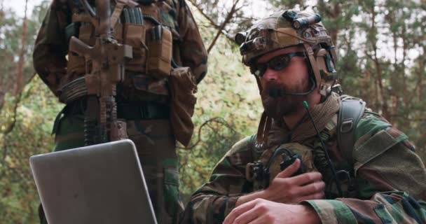 Portret van een bebaarde commandant in een militair uniform in een tactisch vest met een helm op zijn hoofd typend op het toetsenbord van een laptop en meldt een rapport via een militaire radio — Stockvideo