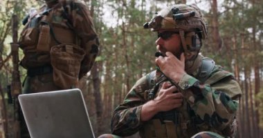 Askeri üniformalı, taktik bir yelek giymiş, başında bir miğferle dizüstü bilgisayarın klavyesine yazı yazan sakallı komutanın portresi ve askeri telsizle bir rapor bildiriyor.