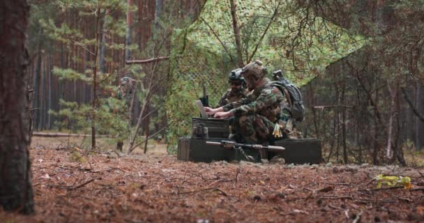 Des soldats barbus en uniforme assis sur des caisses de transport militaire analysent des données sur un ordinateur portable et élaborent des tactiques dans une base forestière temporaire En arrière-plan, vous pouvez voir un soldat protéger la base — Video
