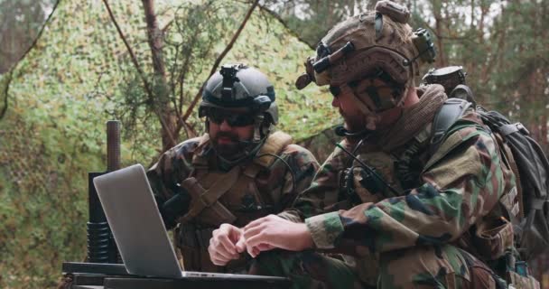 Baard soldaten in uniform zitten op militaire transport kratten analyseren gegevens op een laptop en uit te werken tactiek op een tijdelijke bosbasis Op de achtergrond zie je een soldaat die de basis beschermt — Stockvideo