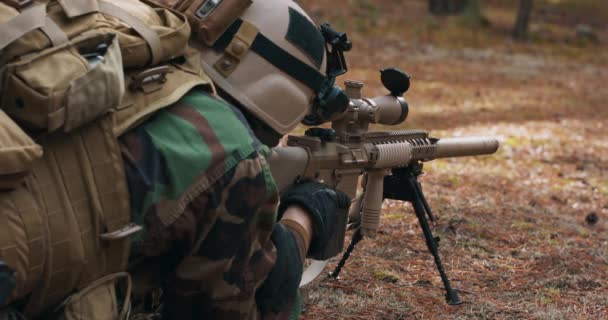 Vousatý voják leží na lesním nosítkách v taktické vojenské uniformě s helmou na hlavě s odstřelovací puškou a míří na nepřítele dalekohledem. — Stock video