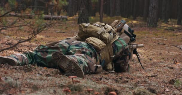 Um soldado barbudo jaz na areia da floresta em um uniforme militar tático com um capacete na cabeça com um rifle sniper e aponta para o inimigo através de um telescópio — Vídeo de Stock