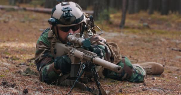 Бородатый солдат лежит на лесном наполнителе в тактической военной форме со шлемом на голове со снайперской винтовкой и открывает метку и целится в врага через прицел — стоковое видео