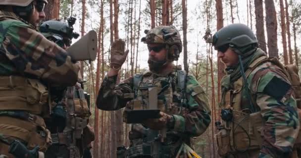 Eine Gruppe bärtiger Soldaten mittleren Alters in Uniformen und taktischen Westen diskutiert den Aktionsplan und bereitet sich auf den Einsatz in einem dichten Wald vor. Der Kommandeur gibt den Soldaten Anweisungen — Stockvideo