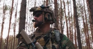Woodland askeri üniformalı, sakallı, orta yaşlı bir askerin portresi ve kafasında kulaklık olan bir miğfer, elinde bir tüfek, kalın çam ağaçlarının arasından etrafa bakıyor. Gün batımında parıltı