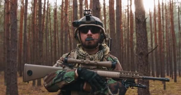 Retrato de un soldado barbudo de mediana edad con uniforme militar y casco de Woodland, con auriculares en la cabeza sosteniendo un rifle. — Vídeo de stock