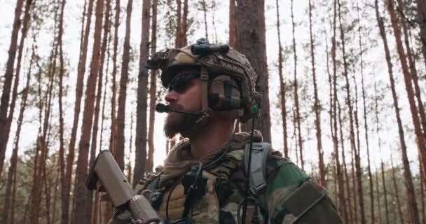 Porträtt av en skäggig medelålders soldat i en militäruniform från skogslandet och en hjälm med hörlurar på huvudet, med ett gevär och med blicken genom den tjocka tallskogen. — Stockvideo
