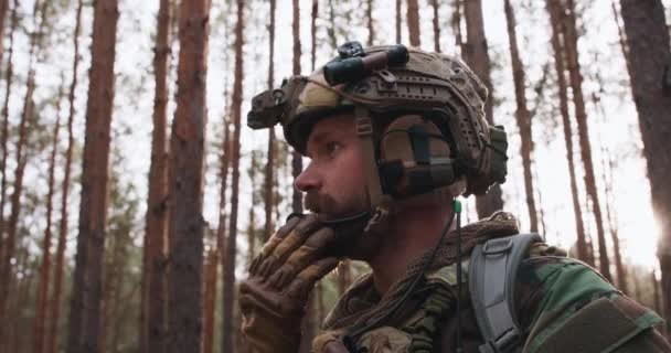 Retrato de un soldado barbudo de mediana edad con un uniforme militar de Woodland y un casco con auriculares en la cabeza que sostiene un rifle y mira a su alrededor a través de un espeso bosque de pinos y habla en la radio. — Vídeo de stock
