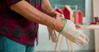 Kot pantolon ve ekose gömlek giyen genç bir marangoz, marangoz dükkanında çalışmaya başlamadan önce beyaz bez koruyucu eldivenler giyer. Arka planda bir çilingir masası ve aletler vardır.