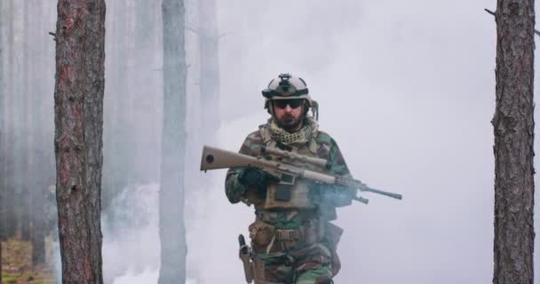 Soldado totalmente equipado con un uniforme de camuflaje que emerge de una nube de humo en medio de un bosque de pinos — Vídeos de Stock