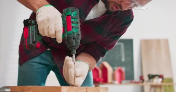 Tukang kayu berjanggut muda terkonsentrasi mengenakan jeans dan kemeja flanel kotak-kotak pengeboran lubang di sepotong kayu dengan bor listrik portabel di bengkel pertukangan — Stok Video