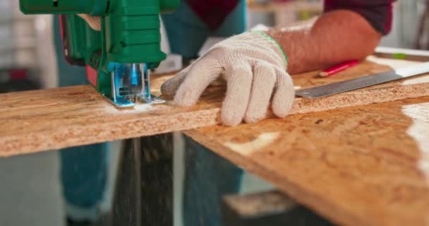 Mladý tesař krájí dřevotřískovou desku řeznou pilou, řemeslníci ruce v ochranných rukavicích. — Stock video