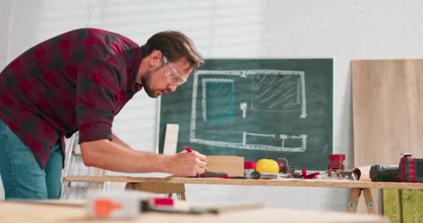 Een hardwerkende professionele timmerman in een flanellen hemd met een potlood terwijl hij een ontwerp analyseert in een timmerwerkplaats Een bebaarde klusjesman met een beschermende bril controleert het ontwerp op een vel — Stockvideo