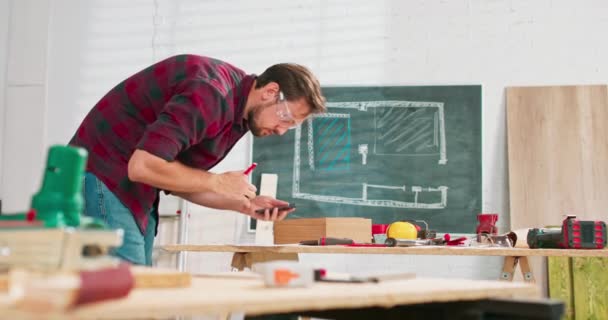 Ένας όμορφος ξυλουργός με φανελένιο πουκάμισο μετράει τις διαστάσεις ενός smartphone σε ένα εργαστήριο ξυλουργικής ένας γενειοφόρος πολυτεχνίτης με προστατευτικά γυαλιά ελέγχει το σχέδιο σε ένα φύλλο χαρτί. — Αρχείο Βίντεο