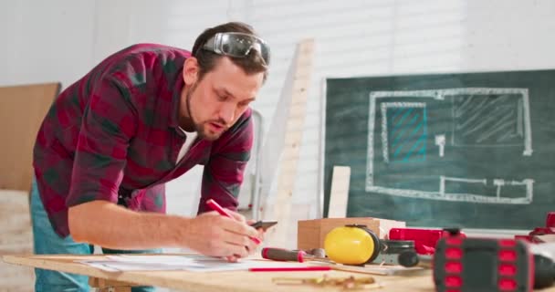 Ένας όμορφος ξυλουργός με φανελένιο πουκάμισο μετράει τις διαστάσεις ενός smartphone σε ένα εργαστήριο ξυλουργικής ένας γενειοφόρος πολυτεχνίτης με προστατευτικά γυαλιά ελέγχει το σχέδιο σε ένα φύλλο χαρτί. — Αρχείο Βίντεο