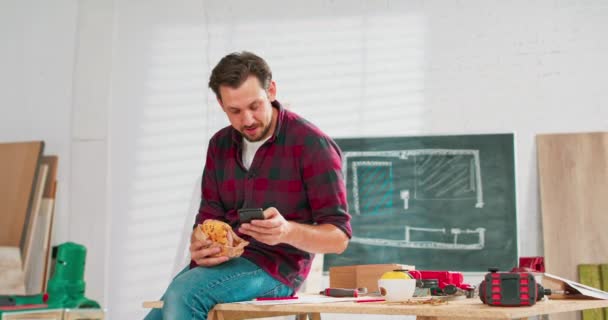 Ένας ξυλουργός χρησιμοποιεί ένα smartphone κατά τη διάρκεια ενός διαλείμματος στη δουλειά Νέος όμορφος τεχνίτης ξεκουράζεται σε ένα εργαστήριο ξυλουργικής Υπάρχει ένα τραπέζι κλειδαράδων και εργαλεία στο παρασκήνιο — Αρχείο Βίντεο