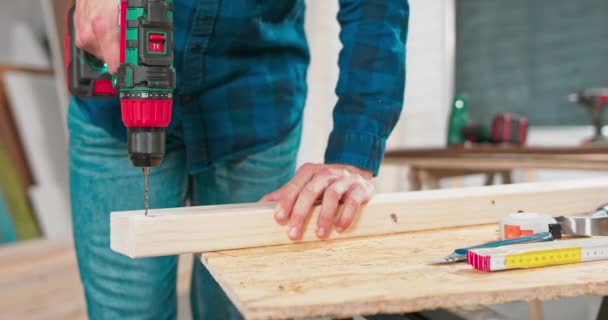 Geconcentreerde jonge bebaarde timmerman draagt jeans en een geruit flanellen hemd boort een gat in een stuk hout met een draagbare elektrische boor in zijn timmermanswerkplaats — Stockvideo