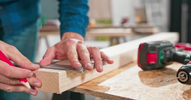 Close-up de um carpinteiro profissional trabalhador segurando uma régua e lápis enquanto mede uma placa em uma oficina de carpintaria. Um entusiasta de bricolage barbudo mede madeira. Há uma mesa de serralheiro e — Vídeo de Stock