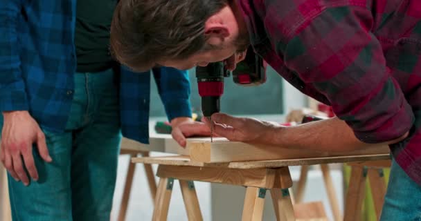 Geconcentreerde jonge bebaarde timmerman draagt jeans en een geruit flanellen hemd boort een gat in een stuk hout met een draagbare elektrische boor in zijn timmermanswerkplaats — Stockvideo