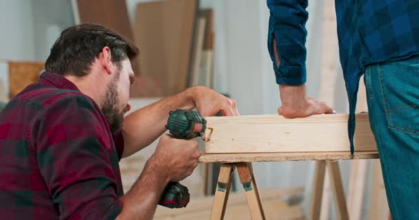 Concentrated young bearded passionate carpenter drives a carpentry screw into a construction beam with a screwdriver in a carpentry workshop — Stock Video