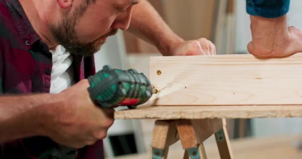 Συμπυκνωμένο νεαρό γενειοφόρο παθιασμένο ξυλουργό οδηγεί μια βίδα ξυλουργικής σε μια δοκό κατασκευής με ένα κατσαβίδι σε ένα εργαστήριο ξυλουργικής — Αρχείο Βίντεο