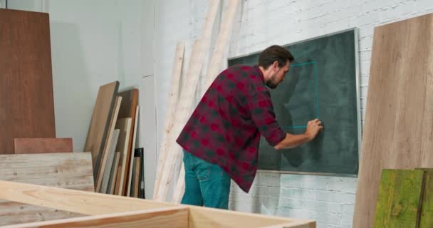 Jonge timmerman bouwt een massief houten muur in een timmerwerkplaats Een meubelmaker tekent een ontwerp van een wandframe op een krijtbord — Stockvideo