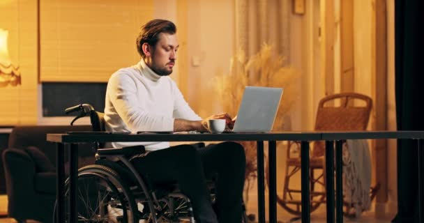 Un joven discapacitado barbudo trabaja en una casa de estilo loft. Se sienta en una silla de ruedas. — Vídeos de Stock