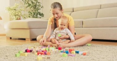 Bir kız çocuğuyla oturma odasındaki halıda oturuyor ve onunla oynuyorlar.