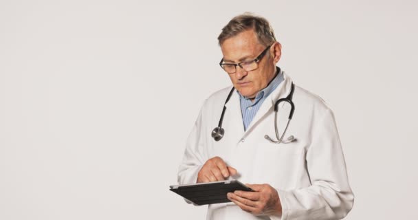 Egy érett orvos, profi, fehér köténybe öltözve, ingben, szemüveggel és sztetoszkóppal körülvéve. — Stock videók