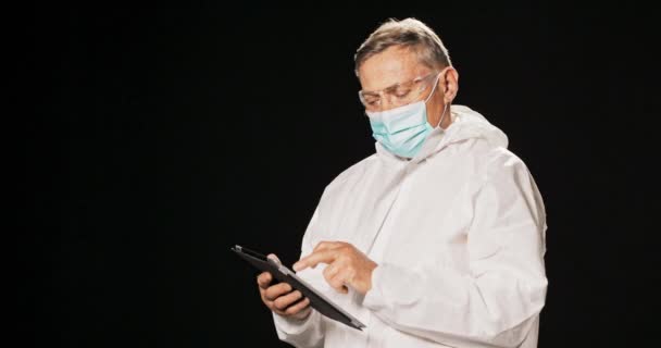 Een oudere man gekleed in beschermende kleding, witte jurk, masker en veiligheidsbril, zoekt naar — Stockvideo