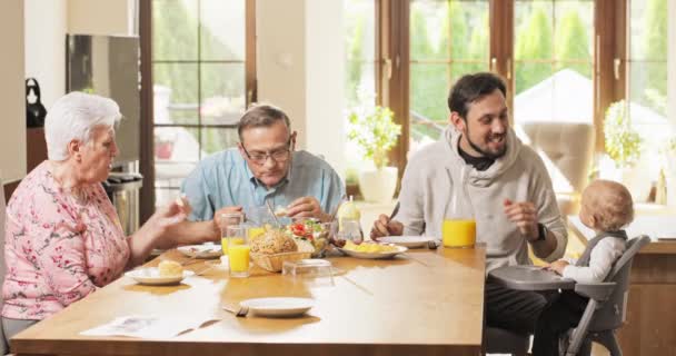 Dziadkowie siedzący z synem i wnukiem przy śniadaniu, rozmawiający, jedzący, człowiek, wspaniały ojciec karmi — Wideo stockowe
