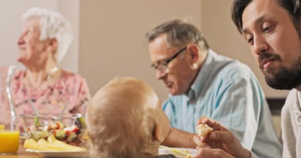 Een brunette man zit aan tafel met zijn ouders en voedt zijn kleine, schattige, — Stockvideo