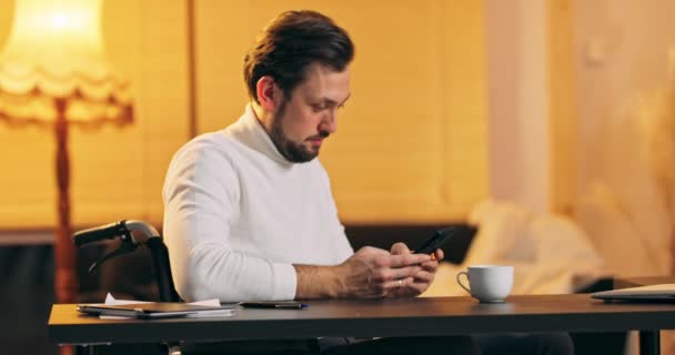 Joven emprendedor en lectura de sillas de ruedas y mensaje de escritura en el teléfono inteligente mientras trabaja en casa — Vídeo de stock