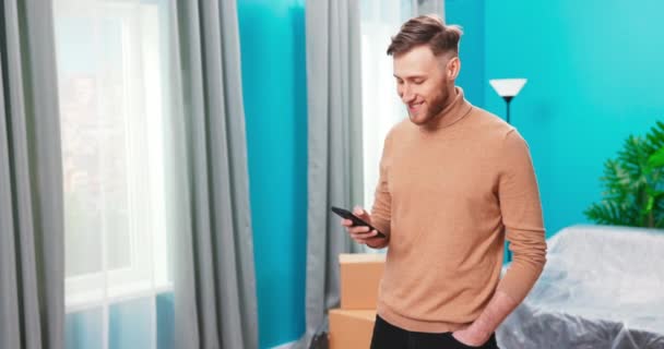 Sonriente hombre milenario de pie utilizando el gadget de almohadilla moderna en la nueva casa de navegación por Internet o de compras, — Vídeos de Stock