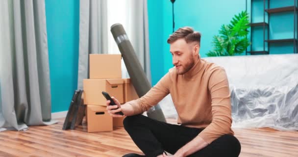 困惑的男人看着智能手机装置，重新安置在新公寓里，搜寻他们公寓的家具 — 图库视频影像