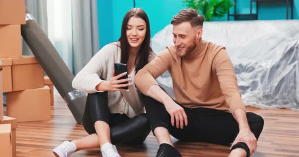 Счастливые молодые кавказские пары используют цифровые смартфоны обсудить идеи обновления интерьера сидеть на — стоковое видео