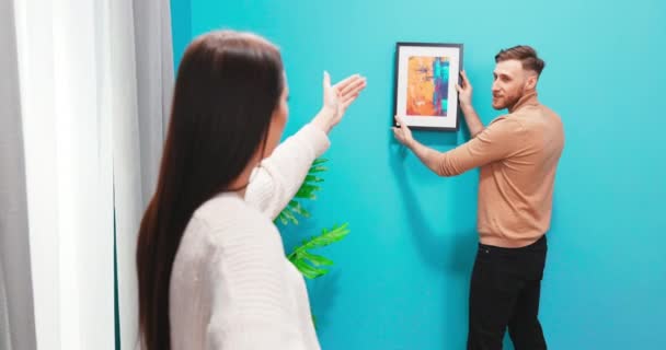 Feliz pareja caucásica se ha mudado a un nuevo apartamento y están decorando el espacio. Mujer diciendo hombre — Vídeos de Stock