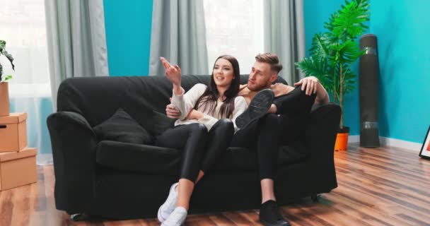 Молода подружня пара домовласників, сидячи на зручному дивані і вирішуючи на кімнатах меблювання або перепланування — стокове відео