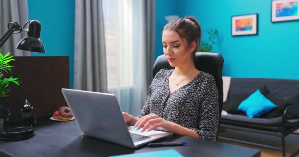 Eine junge hübsche Brünette benutzt einen Laptop, während sie an einem Schreibtisch in einem — Stockvideo