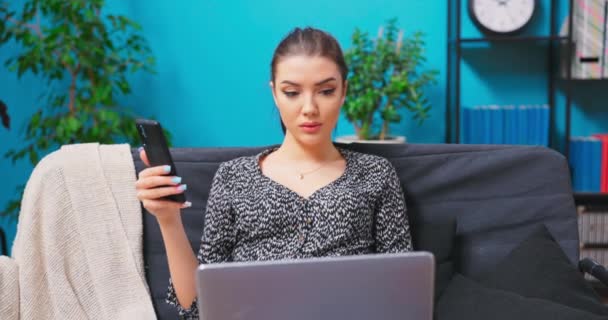 Una chica trabaja en su teléfono inteligente y portátil mientras está sentada en un sofá en un — Vídeos de Stock