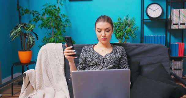 Uma menina trabalha em seu smartphone e laptop enquanto se senta em um sofá em um — Vídeo de Stock