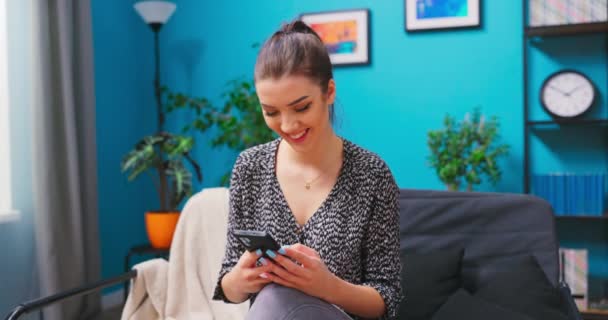 Preety mladá usmívající se žena používá chytrý telefon, zatímco relaxuje ve svém obývacím pokoji. Dívka procházející — Stock video
