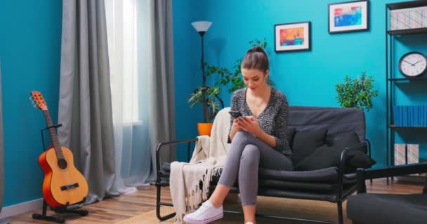 Mladá žena odpočívá na gauči v obýváku a píše sms na smartphonu. A — Stock video