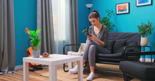 Egy fiatal nő pihen a nappaliban, SMS-ezik az okostelefonján. A... — Stock videók