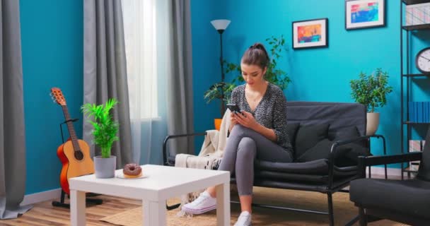 Uma jovem está relaxando no sofá da sala de estar mensagens de texto em seu smartphone. A — Vídeo de Stock
