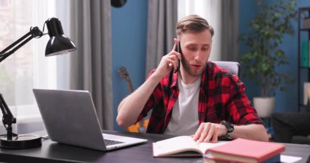 A handsome young man is sitting at his living room desk talking on the phone. — Stock Video