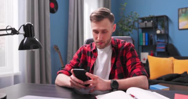 En stilig ung man sitter vid ett skrivbord i ett rum och använder telefonen. — Stockvideo