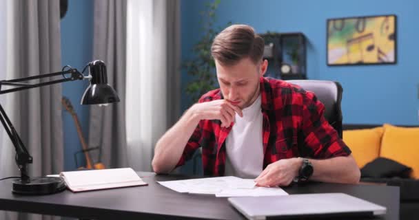 疲れた若い男が自分の部屋の机に座ってアパートをレビューする — ストック動画